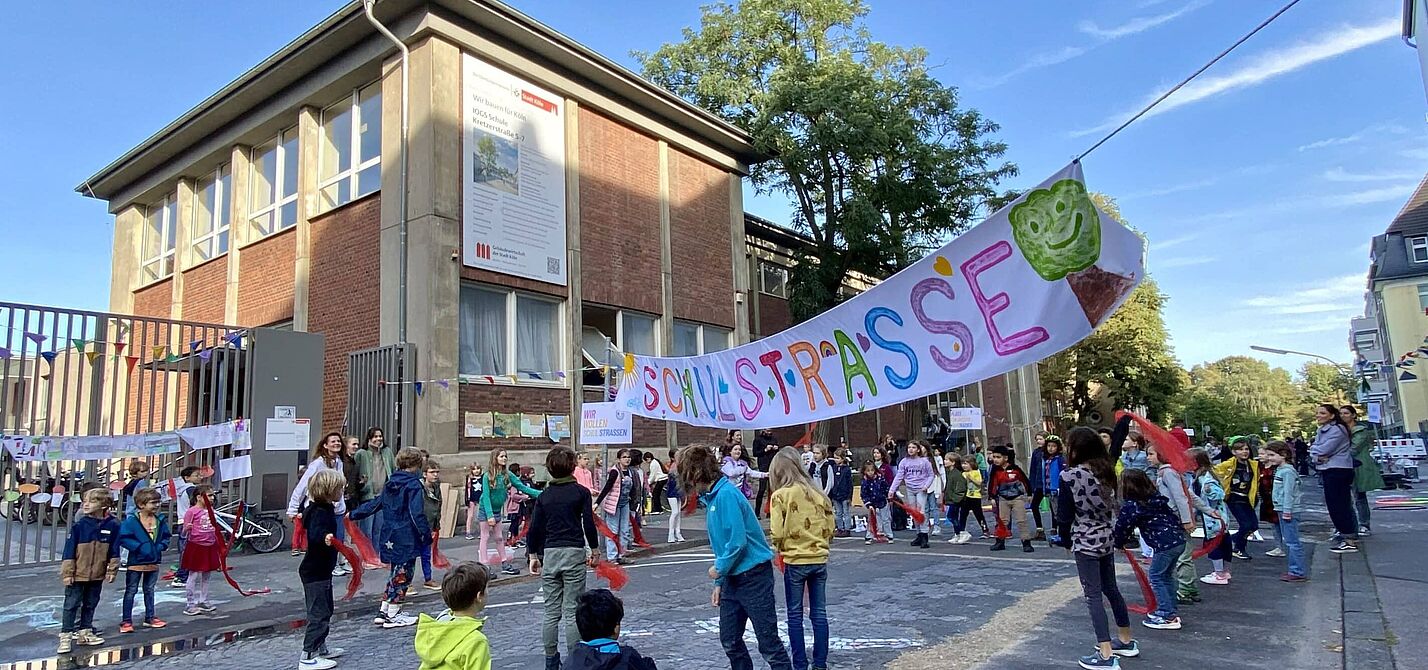 Kinder verschiedenen Alters spielen und bewegen sich auf einer gepflasterten Straße vor einem zweistöckigen Backsteingebäude. Über der Straße hängt ein buntes Banner mit der Aufschrift „Schulstraße“ und einem grünen Baum-Symbol. Im Hintergrund sind Erwachsene zu sehen, die die Aktion begleiten.