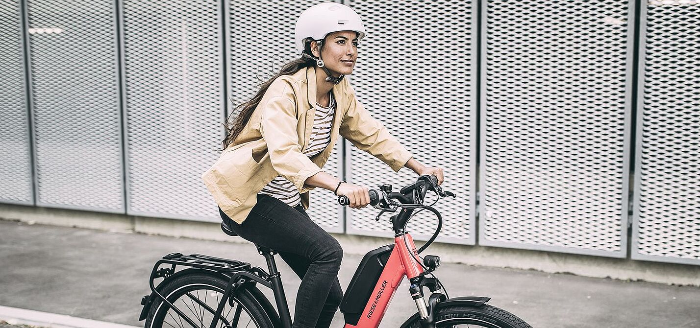 Elektroradtypen Eine Frau fährt auf einem roten Elektrorad auf Asphalt, im Hintergrund graue, gelochte Metallwände.